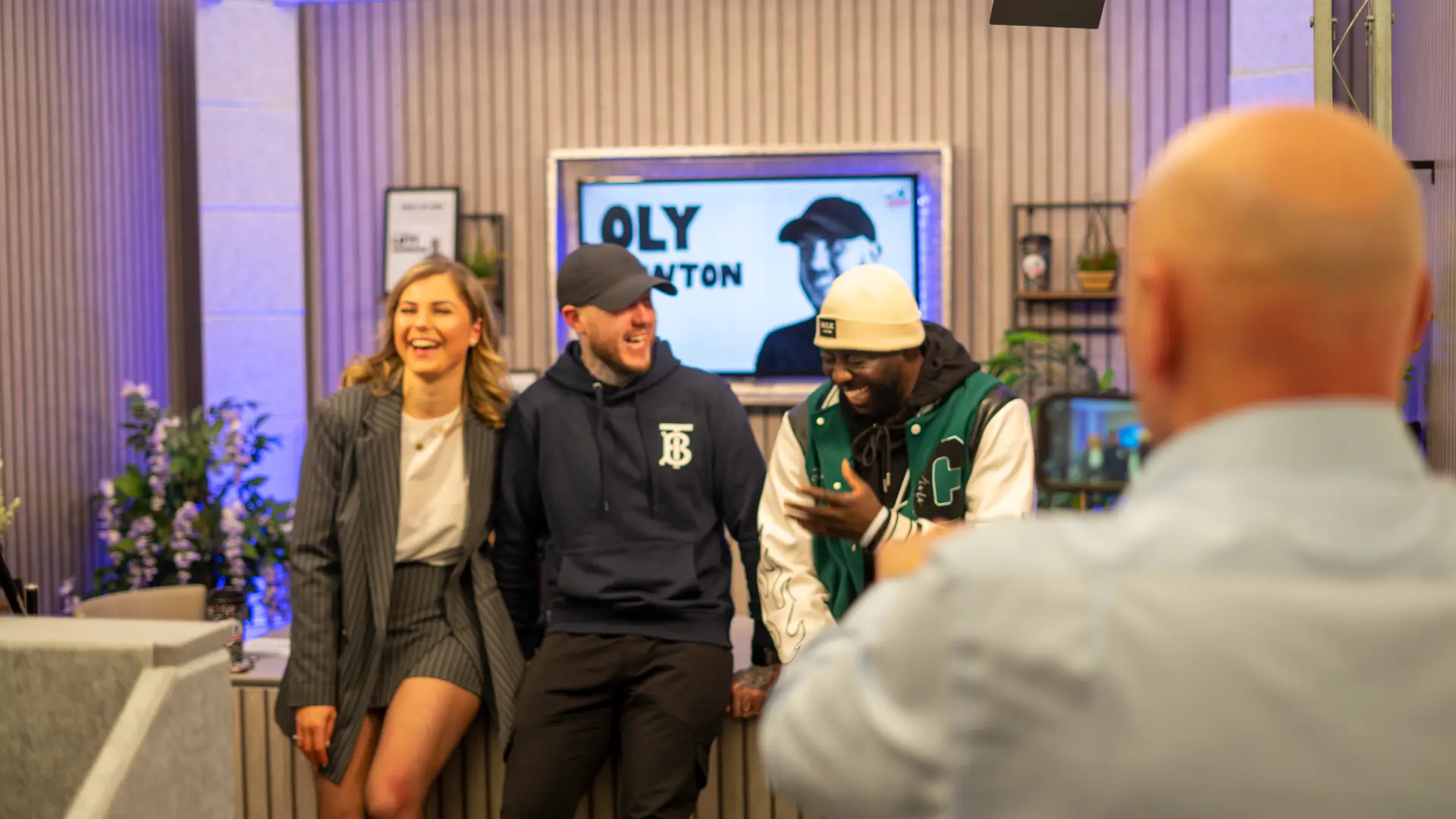 Oly Newton laughing with two fellow guests on a studio set, with his name on the screen behind them