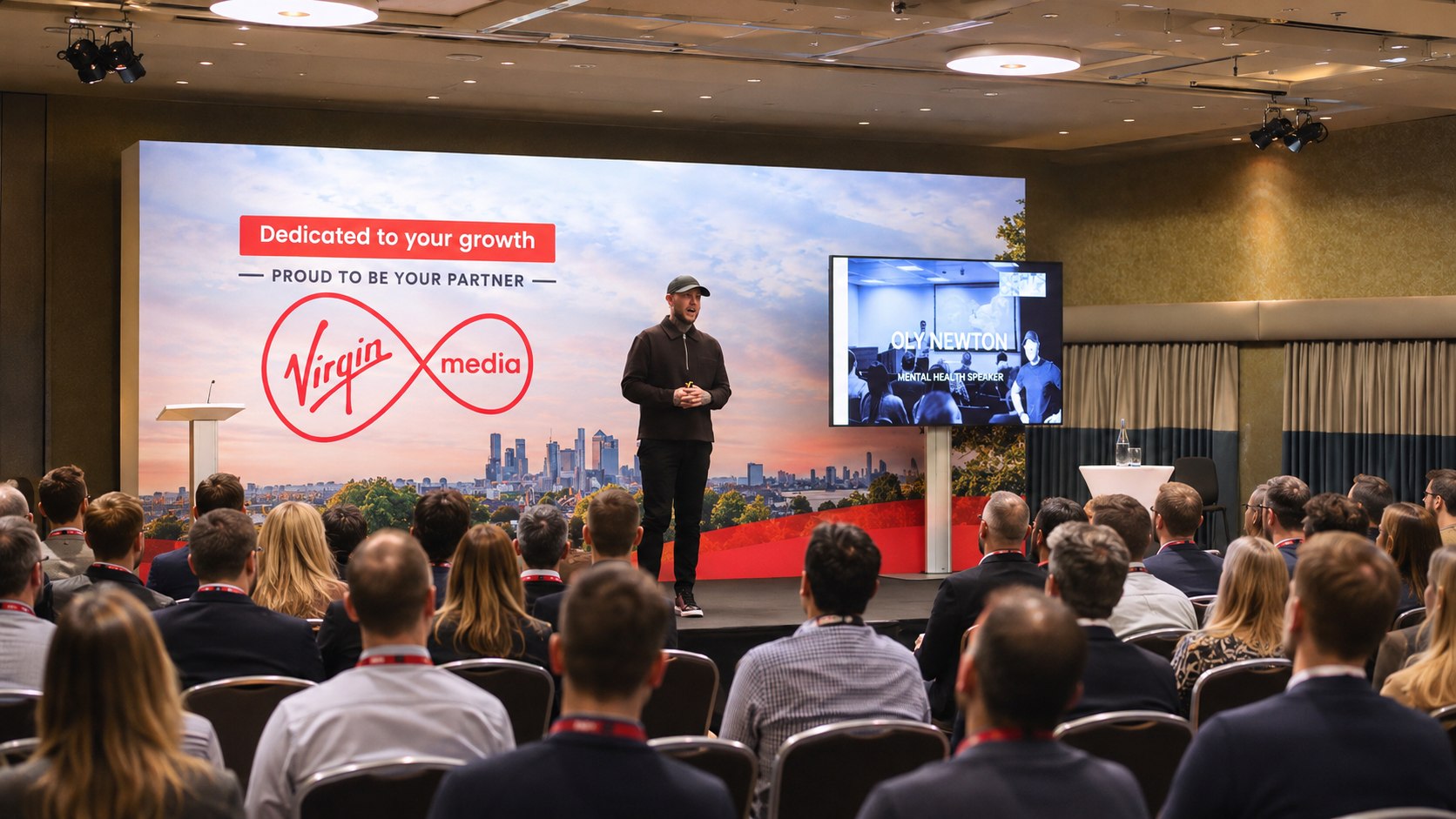Oly Newton speaking on stage at a Virgin Media event, with his name and "mental health speaker" on the screen behind him