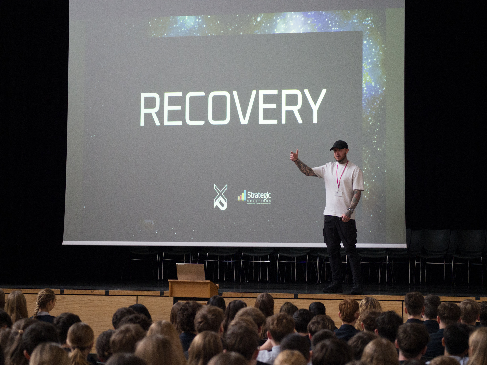 Oly Newton speaking on stage at a school assembly with a Recovery slide behind him and students seated in front