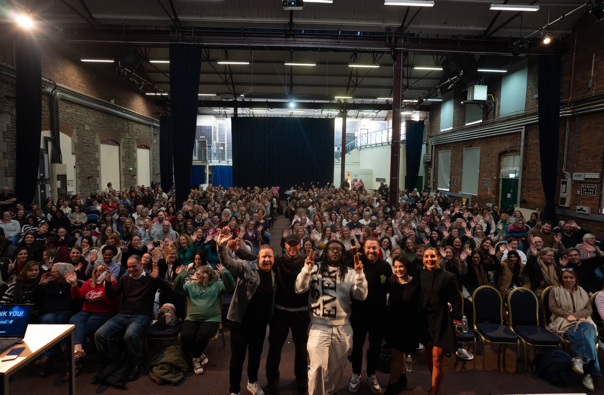 Oly Newton and fellow speakers on stage at the end of a live event, with a full auditorium of people waving behind them