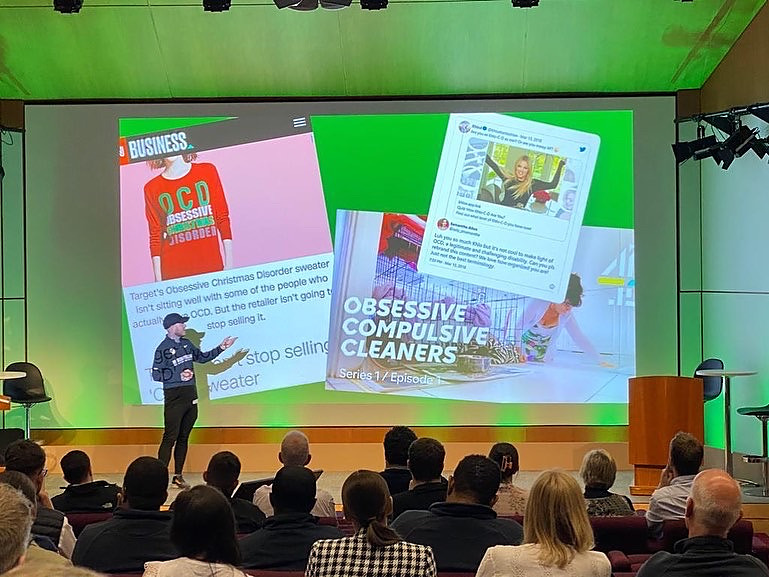 Oly Newton speaking on stage to a full auditorium, with slides on screen behind him showing examples of how OCD is trivialised in the media